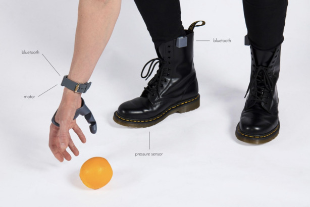 圖中可見，手腕上戴上 Bluetooth 和 Motor，用於無線連接和數據傳輸，以及控制「第三拇指」的動作；而另一 Bluetooth 裝置（在右腳靴上）則用於傳輸裝置在鞋底的 Pressure sensor（壓力感應器）指令。(Photo from Dani Clode Design / The Plasticity Lab)
